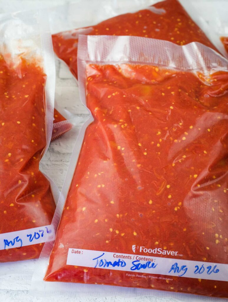 Vacuum-sealed bags filled with tomato sauce, labeled with the date “Aug 2026,” on a white surface.