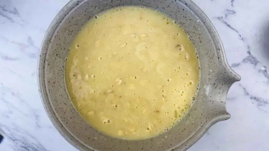 A grey speckled mixing bowl containing a yellow liquid batter with visible oats.