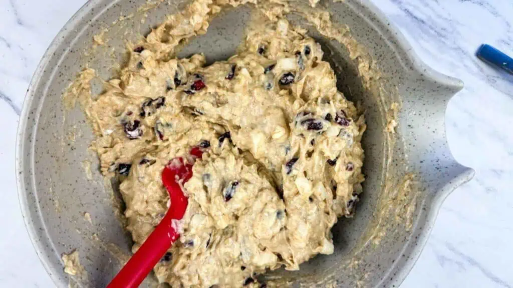 Thick dough with dried cranberries in a ceramic mixing bowl, stirred with a red silicone spatula.