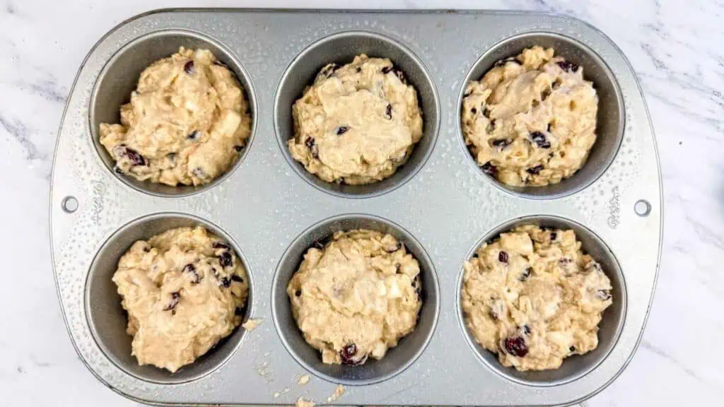 Six muffin cups filled with raw muffin batter containing dried cranberries, set in a metal baking pan.