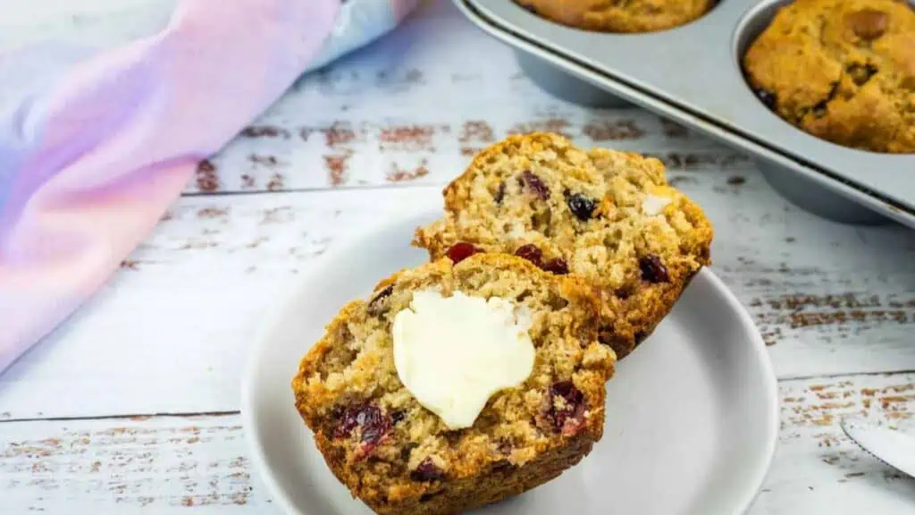A halved cranberry muffin topped with a pat of butter on a small plate, with a muffin tin in the background.