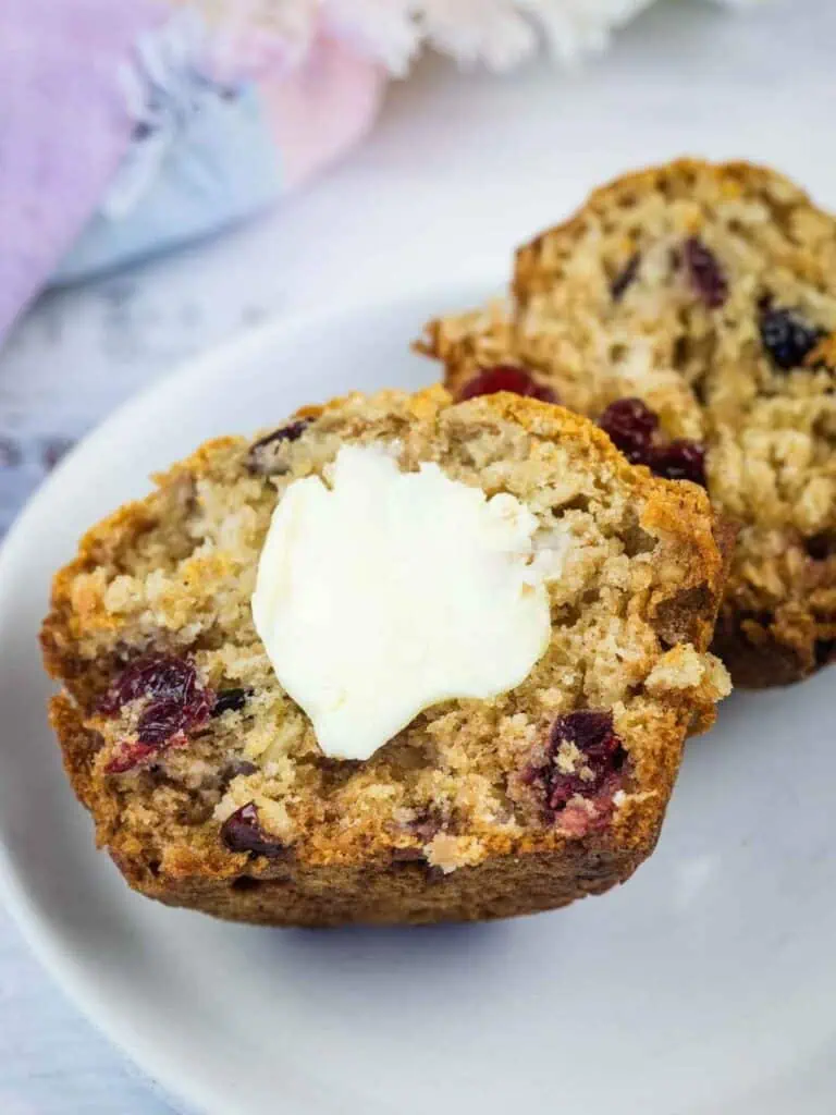 Two halves of a cranberry muffin on a plate, with a pat of butter melting on one half.