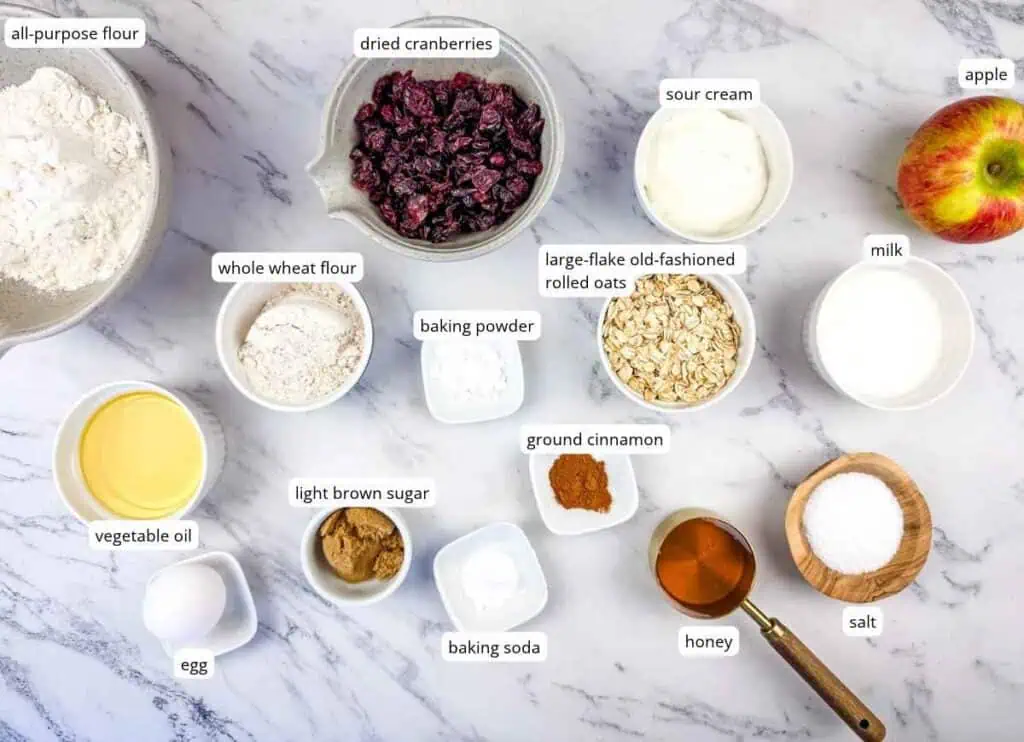 Ingredients for baking, including flour, dried cranberries, sour cream, apple, milk, oats, vegetable oil, egg, brown sugar, baking powder, baking soda, cinnamon, honey, and salt, labeled on a white surface.