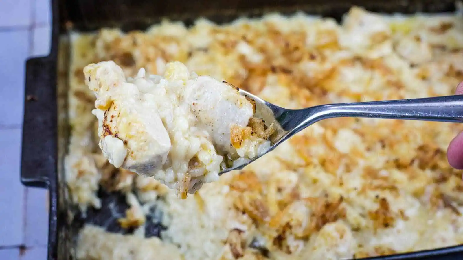 A fork holds a bite of creamy chicken and rice casserole above a baking dish filled with the same casserole.