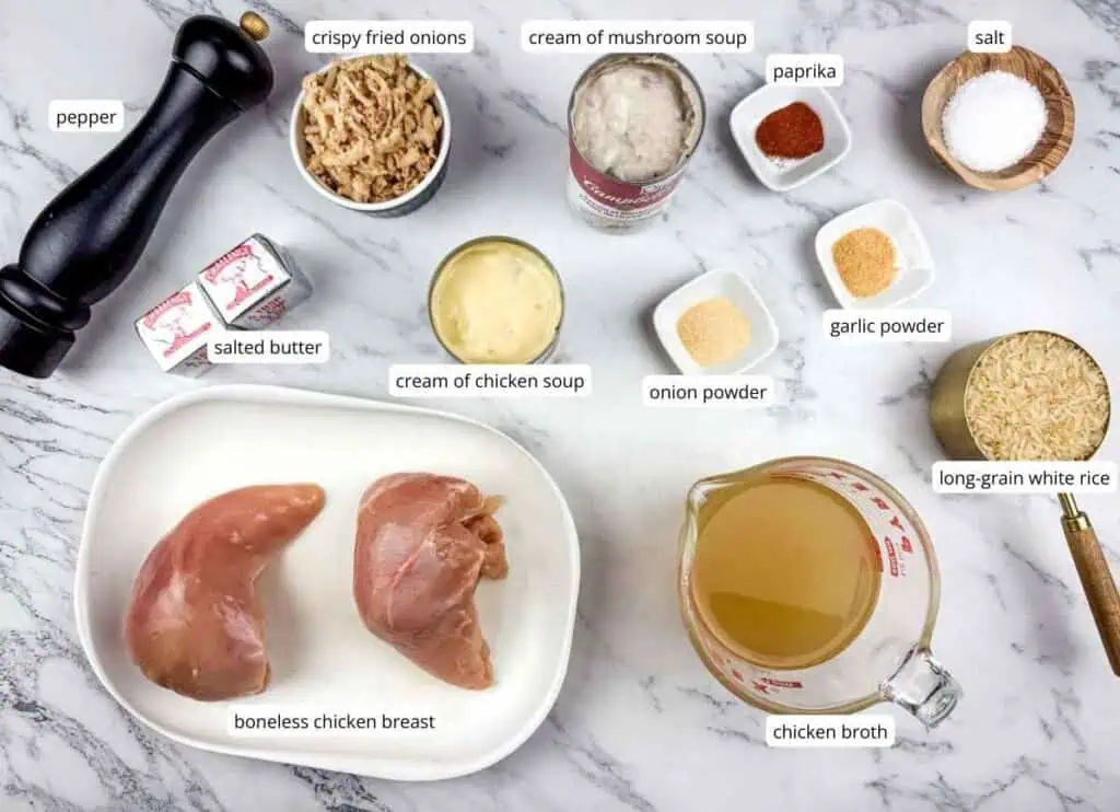 Ingredients for a chicken and rice dish laid out on a marble surface, including chicken breasts, rice, broth, seasonings, soups, butter, and crispy fried onions.