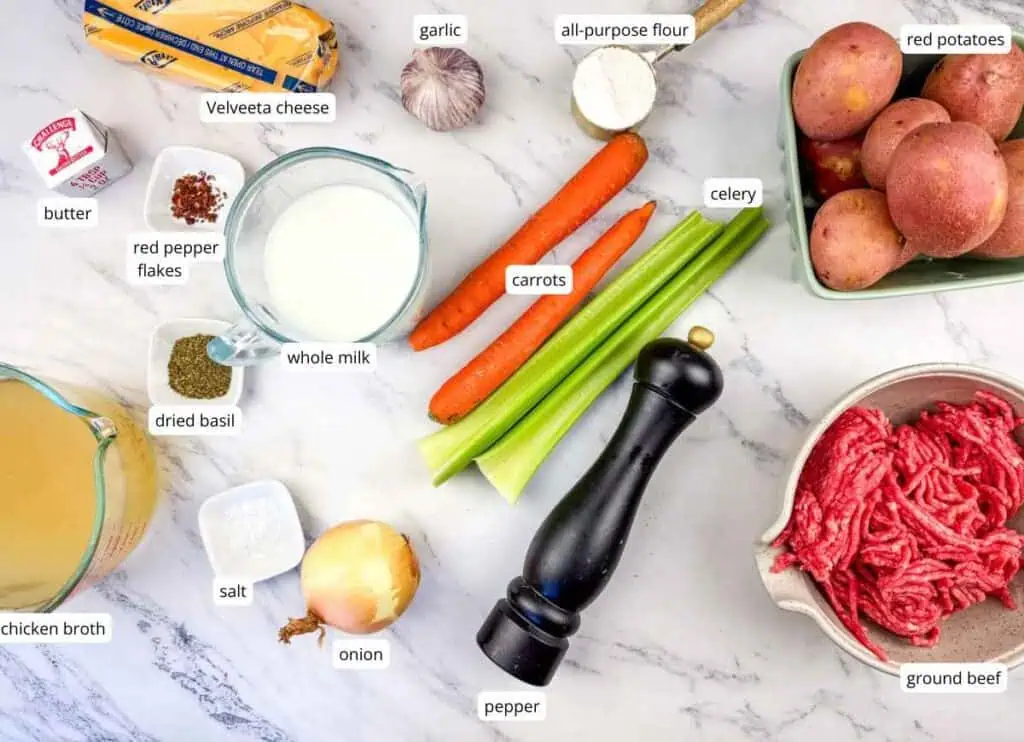 Ingredients for a recipe arranged on a white surface, including red potatoes, ground beef, carrots, celery, onion, garlic, whole milk, butter, Velveeta, chicken broth, flour, and seasonings.