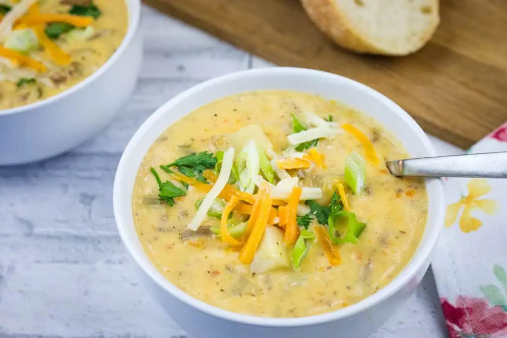 Two white bowls of creamy potato soup topped with shredded cheddar cheese, sliced green onions, and fresh herbs.