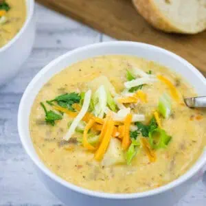 Two white bowls of creamy potato soup topped with shredded cheddar cheese, sliced green onions, and fresh herbs.